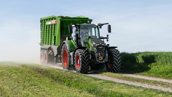 Der grüne Fendt 600 Vario Traktor mit roten Felgen und dem grünen Ladewagen Fendt Tigo fährt auf einer kleinen Feldstraße mit einer grünen Wiese im Vorder- und Hintergrund.