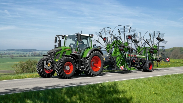 Der grüne Traktor Fendt 600 Vario mit roten Felgen fährt mit dem grünen Fendt Former auf einer grauen Straße. Im Hintergrund ist eine große grüne Wiese und ein blauer Himmel.