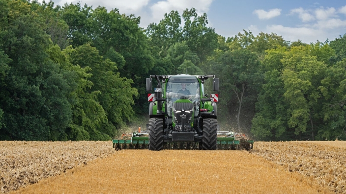 Ein grüner Fendt 620 Vario fährt mit einer Scheibenegge über ein Stoppelfeld im Hintergrund ein Wald mit grünen Bäumen.