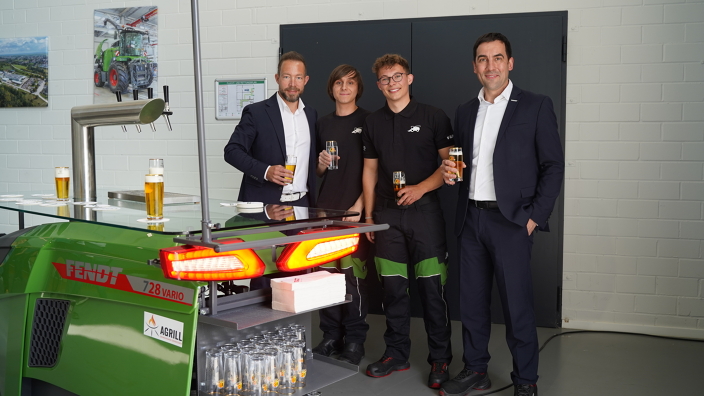 4 Männer, davon 2 Auszubildende (Mitte), stehen mit Kaltgetränken hinter einer grünen Fendt Zapfanlage mit leuchtenden Rücklichtern, einer Glasplatte als Abstellmöglichkeit und Gläserregalen.