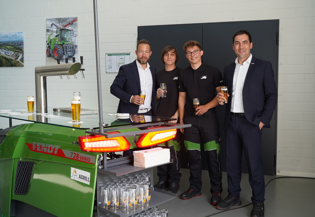 4 Männer, davon 2 Auszubildende (Mitte), stehen mit Kaltgetränken hinter einer grünen Fendt Zapfanlage mit leuchtenden Rücklichtern, einer Glasplatte als Abstellmöglichkeit und Gläserregalen.