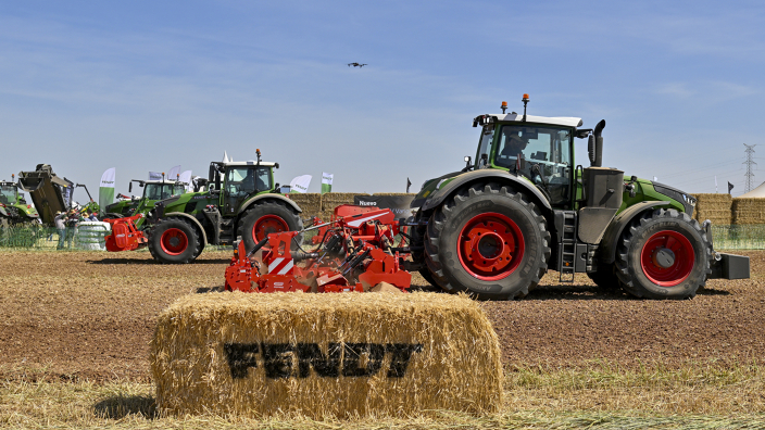 Ein Strohballen mit Fendt Logo im Vordergrund, dahinter fahren zwei grüne Fendt Traktoren mit Anbaugeräten über einem Acker