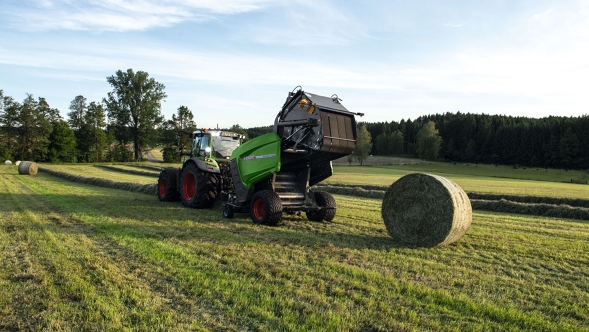 Die Fendt Rotana 160 V Xtra fährt auf einem Feld und presst Rundballen. Im Hintergrund sind ein Wald und der blaue Himmel zu sehen.