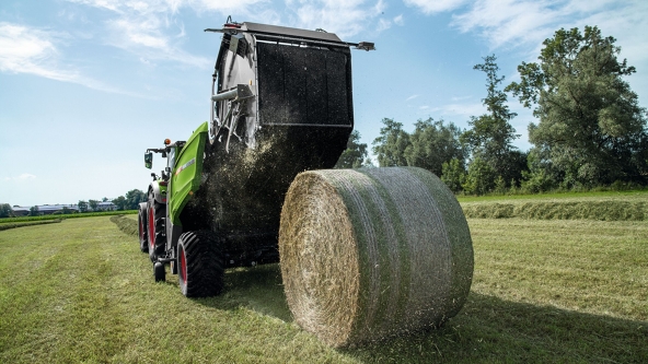 Die Fendt Rotana fährt auf einem Feld und hat gerade einen fertigen Heuballen ausgegeben. Im Hintergrund sind Bäume und der blaue Himmel zu sehen.