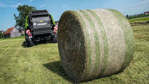 Im Vordergrund steht der fertige Heuballen, während die Fendt Rotana das Feld weiter entlangfährt. Im Hintergrund sind Häuser zu erkennen.