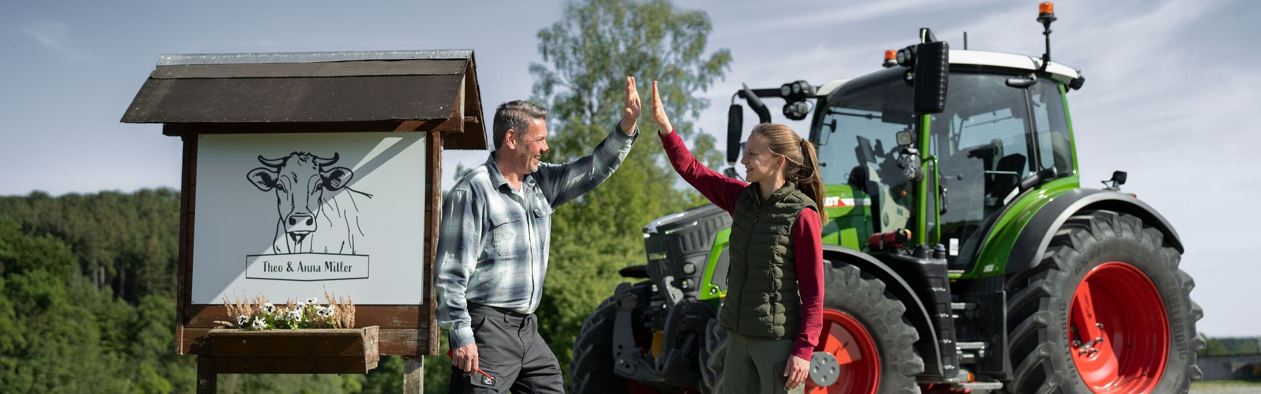 Ein Mann und eine Frau geben sich auf einem Feldweg vor einem Fendt 500 Vario Traktor ein High Five, neben ihnen steht ein Hofschild mit einer gezeichneten Kuh und der Aufschrift „Theo und Anna Miller“