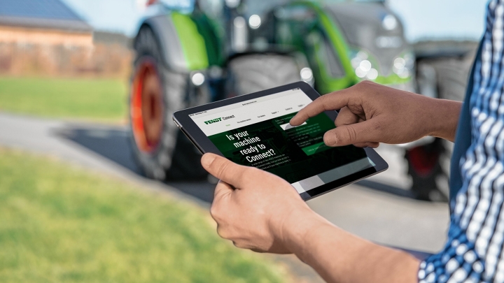 Nahaufnahme eines Tablets in der Hand eines Landwirts, der per Fendt Connect seine zentralen Maschinendaten prüft. Im Hintergrund sieht man leicht verschwommen einen grünen Traktor der Marke Fendt.