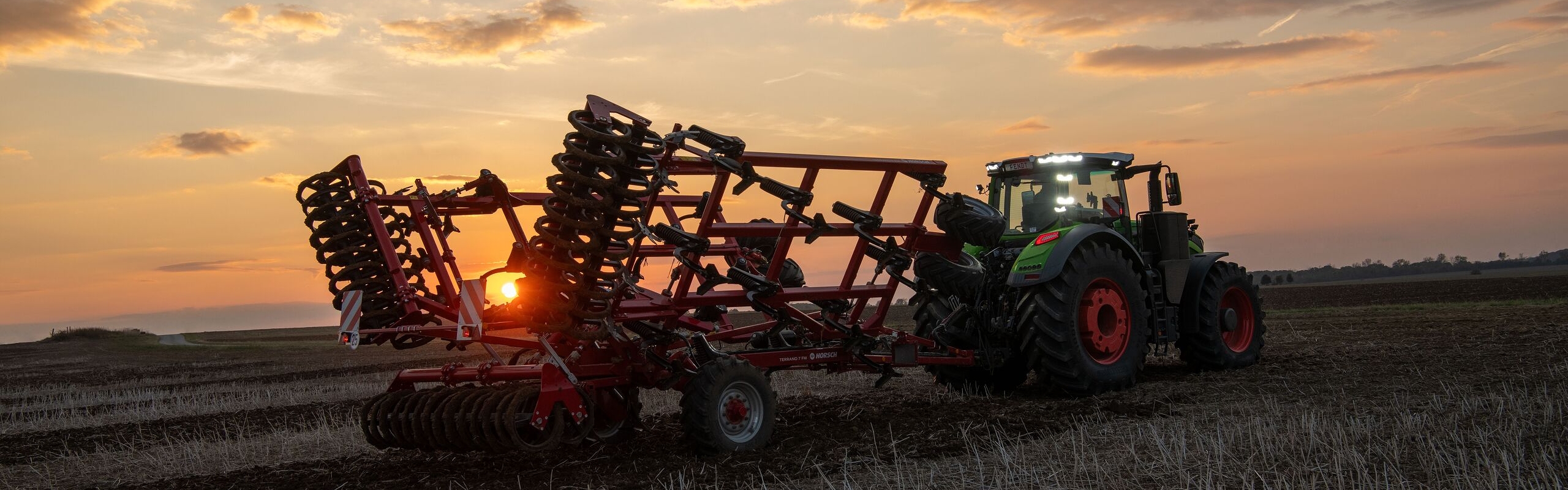 Im Sonnenuntergang fährt ein Fendt 1000 Vario mit Anbaugerät über ein Feld