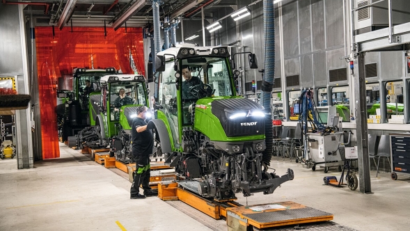 In einer hellen Werkshalle stehen mehrere grüne, unfertige Fendt-Traktoren in einer Reihe. In den Traktor-Kabinen sitzen Mitarbeiter. Vorne steht zudem ein Mitarbeiter, der etwas außen montiert. Im Hintergrund sieht man diverse Werkzeuge und Maschinen.