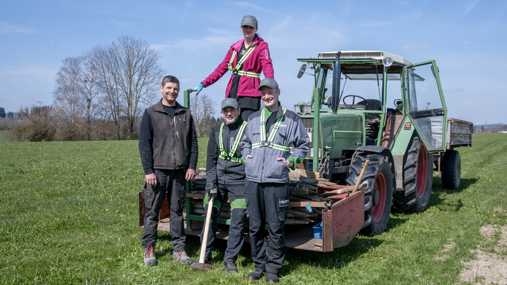 Fendt Mitarbeitende am Aktionstag #TEAMFENDT mit Landwirt Martin Reiter 4 Personen, davon eine Frau, die auf den aufgeladenen Holzpfählen auf dem Fendt Traktor steht. Die 3 Männer stehen vor dem grünen Fendt Traktor.