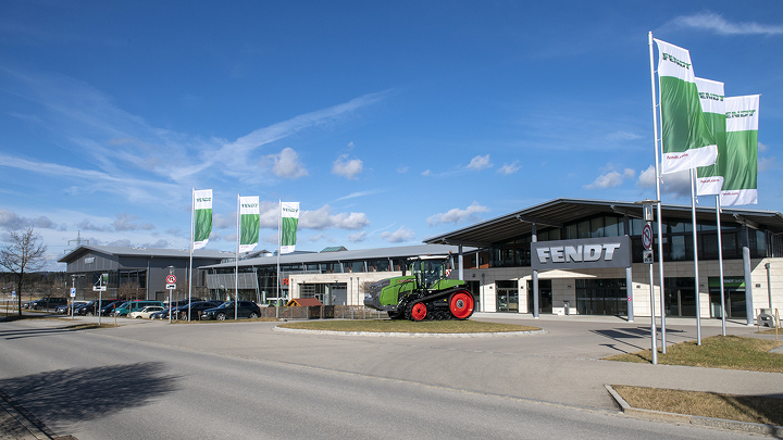 Rechts ein großes Gebäude mit einem Fendt Schild, links daneben steht ein Fendt Traktor und im Vordergrund weiß/grüne Fendt Fahnen.