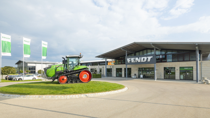 Rechts ein großes Gebäude mit einem Fendt Schild, links daneben steht ein Fendt Traktor mit weiß/grüne Fendt Fahnen.