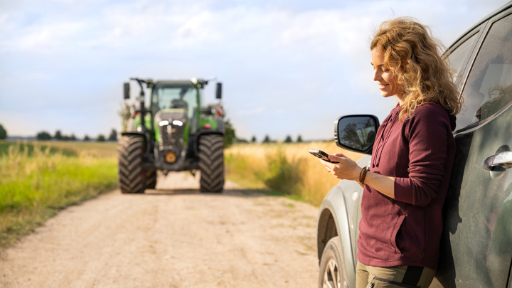 Ein Landwirtin steht neben einem Auto und schaut auf Ihr Smartphone, im Hintergrund ist ein Fendt Traktor zu sehen.