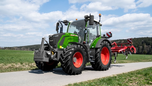 Ein Fendt 300 Vario Gen5 Traktor fährt bei Tageslicht mit angebautem roten Anbaugerät über eine schmale Straße durch eine ländliche Landschaft