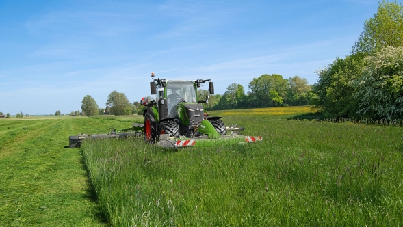 Ein Fendt 300 Vario Gen5 Traktor mäht mit einem Front- und Heckmähwerk eine dichte grüne Wiese an einem sonnigen Frühlingstag