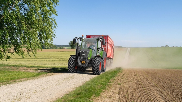 Ein Fendt 300 Vario Gen5 Traktor fährt mit einem roten Anhänger auf einem Schotterweg zwischen Feldern und wirbelt dabei Staub auf bei strahlendem Sonnenschein