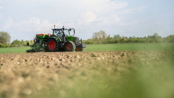 Ein Fendt 300 Vario Gen5 Traktor fährt über ein offenes Feld und bearbeitet den Boden bei klarem Himmel mit weißen Wolken im Hintergrund.