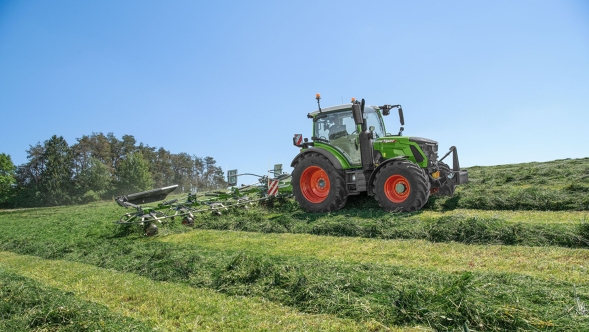 Ein Fendt 300 Vario Gen5 Traktor fährt über eine gemähte Wiese und schwadet das Gras in gleichmäßige Bahnen bei strahlendem Sonnenschein