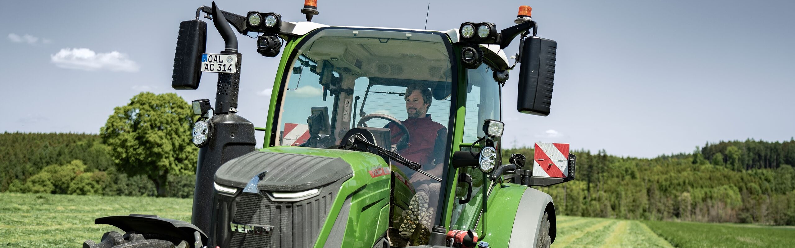 Ein Fendt 300 Vario Gen5 Traktor mäht mit einem Frontmähwerk eine saftig grüne Wiese bei Sonnenschein, der Fahrer sitzt konzentriert in der Kabine
