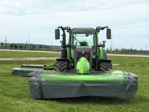 Frontalansicht eines grünen Fendt Traktors mit angebautem Frontmähwerk auf einer ebenen Wiese. Das Mähwerk ist vollständig ausgeklappt, mit grauen Schutztüchern und grün lackierter Verkleidung. Das System ermöglicht eine Querschwenkung von +/- 5° für idea