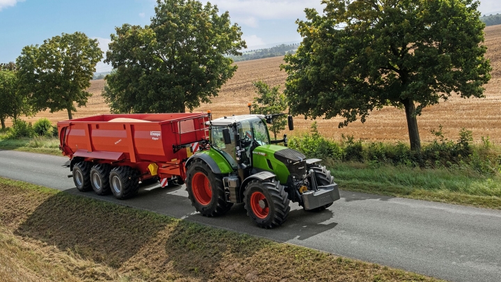 Der grüne Fendt 700 Vario fährt mit einem roten Anhänger auf einer Landstraße. Im Hintergrund sind Bäume, ein Feld und der blaue Himmel zu sehen.
