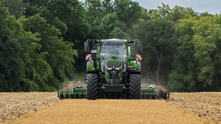 Fendt 600 Vario Der grüne Fendt 600 Vario ist im Einsatz mit einem Anbaugerät auf einem braunen Feld. Im Hintergrund ist ein dunkelgrüner Wald und ein wolkenbehangener Himmel.