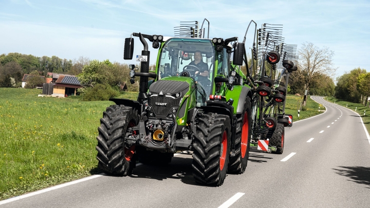 Der grüne Fendt 600 Vario fährt mit einem Fendt Former auf einer Landstraße, umgeben von grünen Feldern und Bäumen. Im Hintergrund sieht man den leicht bewölkten Himmel.