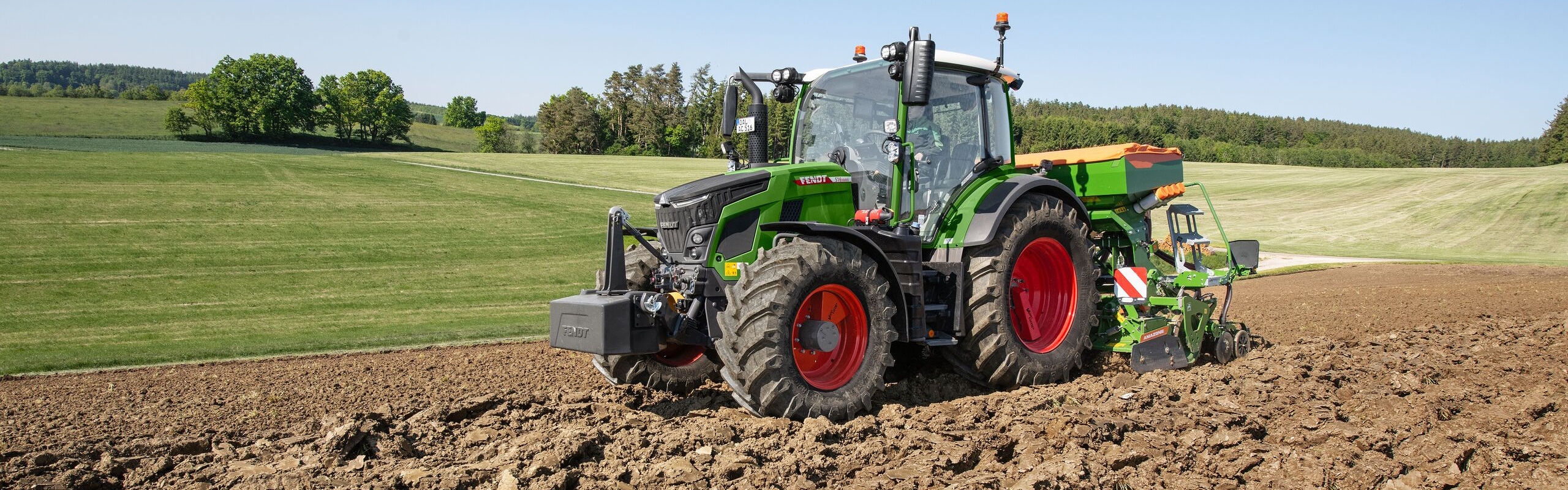 Ein Fendt 500 Vario Traktor mit angebauter Sätechnik arbeitet auf einem frisch gepflügten Acker bei Sonnenschein, im Hintergrund grüne Wiesen und sanfte Hügel