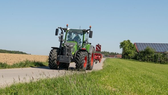 Ein Fendt 500 Vario Traktor mit angebautem Arbeitsgerät fährt bei Sonnenschein über eine schmale Landstraße zwischen Feldern und Wiesen, im Hintergrund ein Gebäude mit Solaranlage