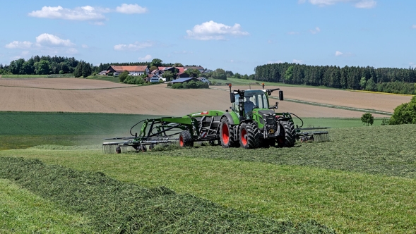 Fendt 500 Vario Gen4 mit Schwader beim Zusammenlegen von gemähtem Gras auf einer hügeligen Wiese