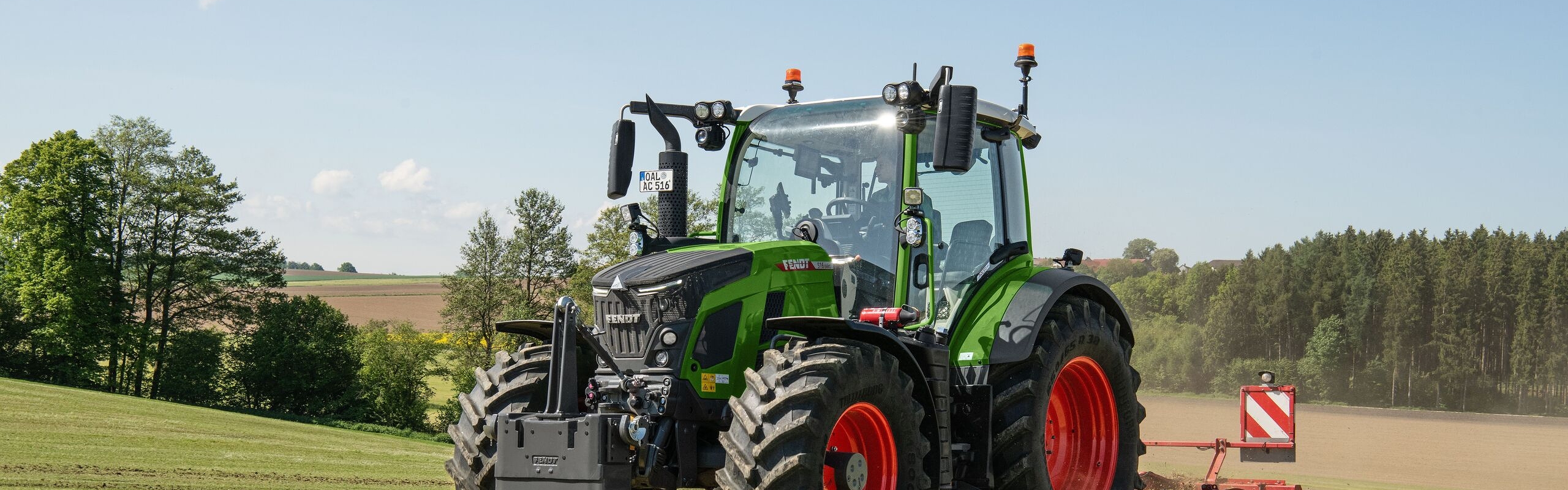 Ein Fendt 500 Vario Gen4 Traktor in grün mit roten Felgen fährt auf einer hellgrünen Wiese. Hinten ist ein rotes Bodenbearbeitungsgerät mit Warntafel angehängt. Im Hintergrund sind Felder, ein Waldrand und blauer Himmel mit wenigen Wolken zu sehen.
