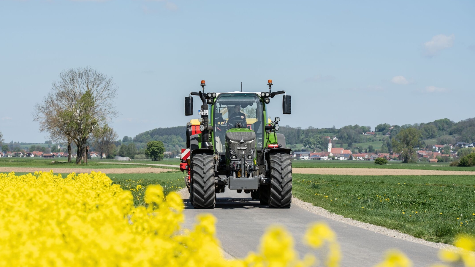 Ein Fendt 500 Vario Traktor fährt auf einer schmalen Straße durch die Landschaft, im Vordergrund blüht ein leuchtend gelbes Rapsfeld, im Hintergrund sind Felder und ein Dorf zu sehen