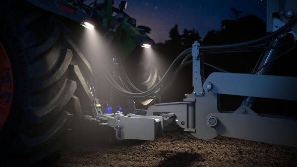 Bei Nachtaufnahme zeigt die Beleuchtung des Fendt 500 Vario Gen4 gezielt den hinteren Anbauraum. Zwei helle LED-Strahler oberhalb des großen linken Hinterreifens leuchten auf die Unterlenker, Hydraulikanschlüsse und den trockenen, braunen Boden. Im Hintergrund sind dunkle Baumumrisse vor einem sternklaren Himmel zu erkennen.