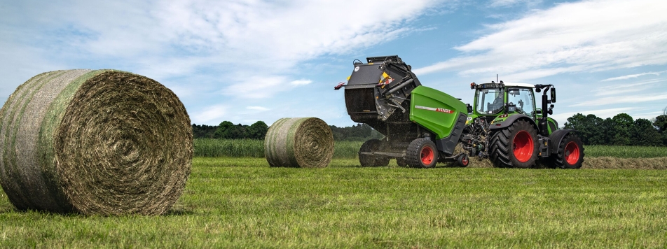 Auf einem Feld fährt ein Fendt Traktor mit angehängter Fendt Rundballenpresse Rotana 160 V am Horizont, davor zwei fertig gepresse Rundballen