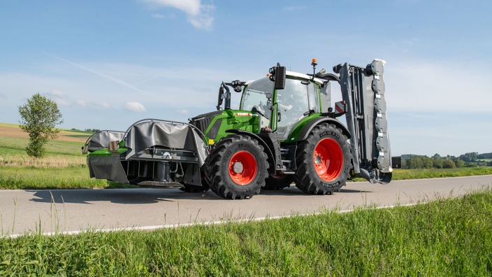 Ein grüner Fendt 300 Vario fährt mit einem Fendt Slicer Front- und Heckmähwerk auf einer Straße.