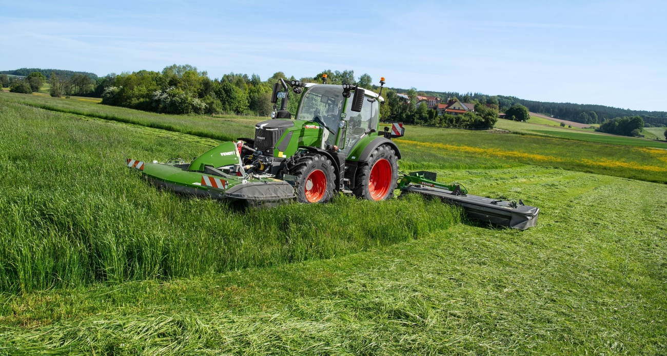 Ein grüner Fendt 300 Vario mäht mit einem Fendt Slicer Front- und Heckmähwerk eine hügelige Wiese.