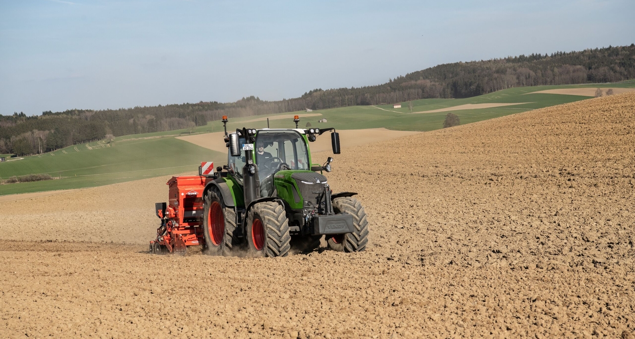 Ein grüner Fendt 300 Vario fährt mit einer roten Drillmaschine und Frontgewicht über ein Feld während der Aussaat.