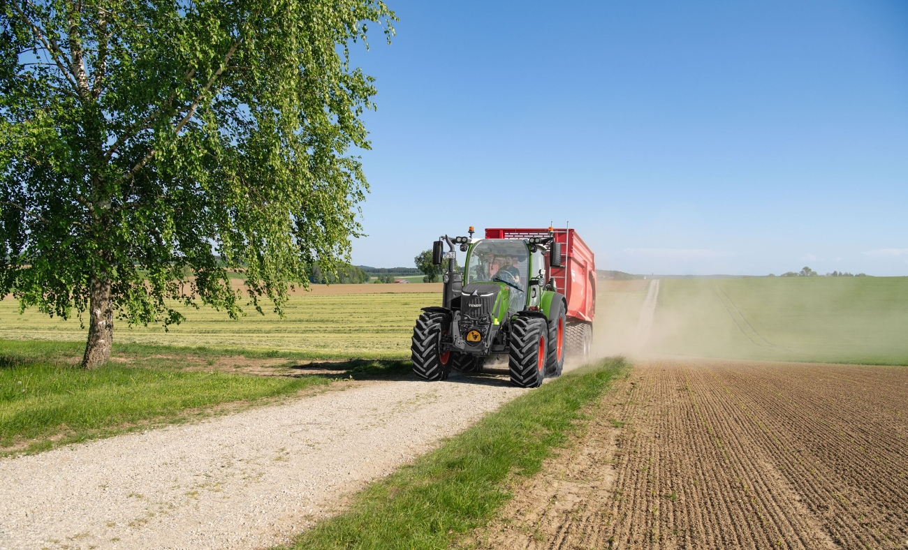 Ein grüner Fendt 300 Vario fährt mit einem roten Anhänger einen Feldweg hinauf.