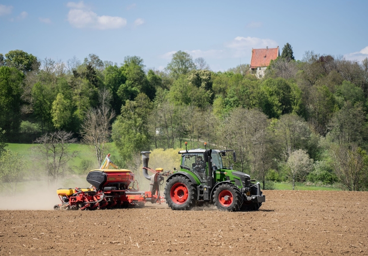 Ein Fendt 500 Vario im Einsatz mit einer Einzelkornsämaschine Ein grüner Fendt 500 Vario fährt im Gespann mit einer Einzelkornsämaschine über einen Acker, im Hintergrund ein Wald mit einem Haus.
