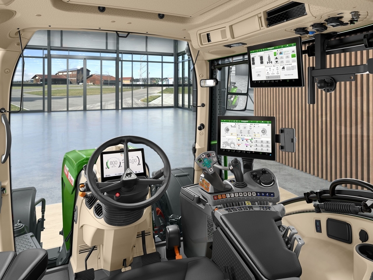 Kabine des Fendt 500 Vario Eine Innenaufnahme der Traktorkabine aus Sicht des Fahrers mit Lenkrad, FendtONE Bedienarmlehne mit Joystick und Terminal sowie einem zusätzlichen Terminal im Dach. Durch die Frontscheibe sieht man eine Halle mit großem Fenster und einen Hof hinter dem Fe