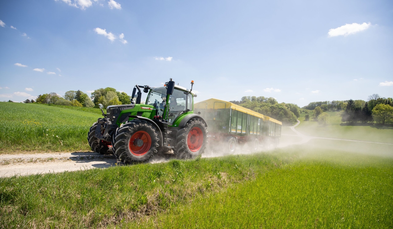 Ein grüner Fendt 500 Vario zieht zwei grüne Anhänger mit gelber Folienabdeckung über einen Feldweg und wirbelt Staub auf.