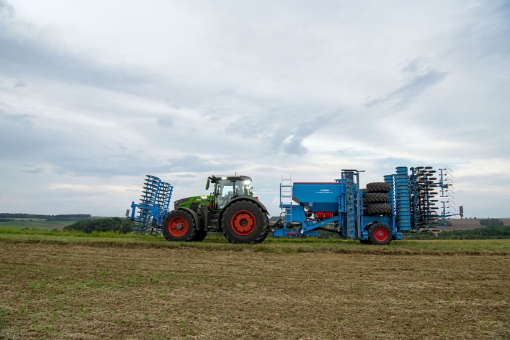 Fendt 800 Vario im Transport mit Frontpacker und einer Einzelkorndrille Ein grüner Fendt 800 Vario fährt im Gespann mit einem Frontpacker und einer blauen Einzelkorndrille von Lemken im Heck über einen Feldweg.