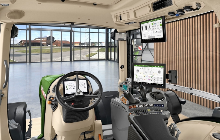 Innenraum der Fendt Kabine Eine Innenaufnahme der Traktorkabine aus Sicht des Fahrers mit Lenkrad, FendtONE Bedienarmlehne mit Joystick und Terminal sowie einen zusätzlichen Terminal im Dach. Durch die Frontscheibe sieht man eine Halle mit großem Fenster und einen Hof hinter dem Fe