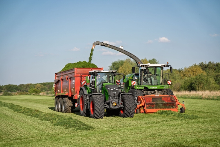Ein Fendt 800 Vario mit einem Fendt Katana Feldhäcksler Ein grüner Fendt 800 Vario fährt im Gespann mit einem Anhänger neben einem Fendt Katana Feldhäcksler über das Grünland