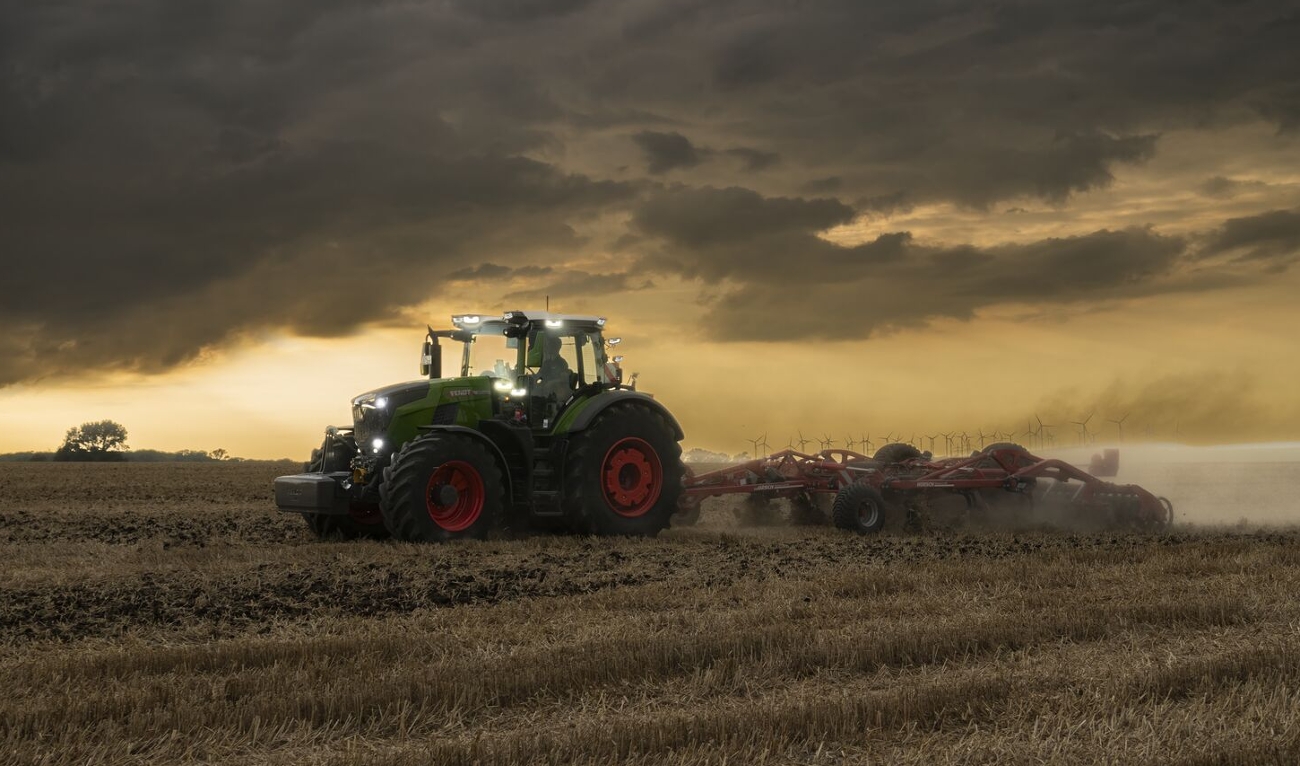 Ein grüner Fendt 800 Vario fährt mit einem Grubber auf einem Stoppelfeld in den Abendstunden und wirbelt Staub auf, im Hintergrund sind einige Windräder zu sehen.