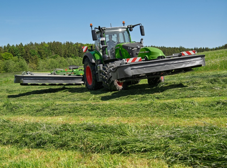 : Ein Fendt 724 Vario Gen7.1 wendet mit Mähwerk Ein grüner Fendt 724 Vario Gen7.1 wendet mit angehobenen Fendt Slicer Front- und Heckmähwerken auf einer Wiese.