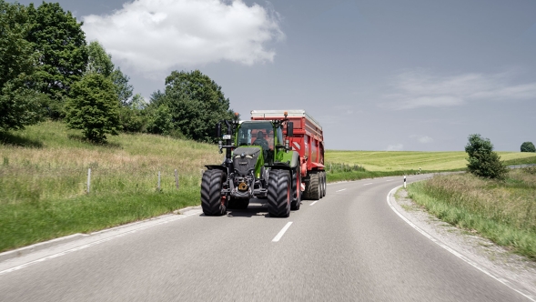 Fendt 700 Vario Gen7.1 mit Anhänger auf kurviger Landstraße bei sommerlichem Wetter. Fendt 700 Vario Gen7.1 mit rotem Anhänger auf Landstraße zwischen grünen Wiesen und Bäumen bei Sonnenschein.