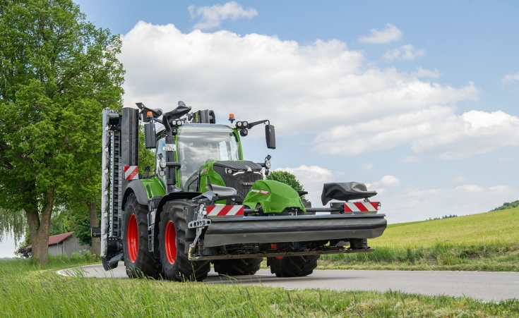 Fendt 724 Vario Gen7.1 transportiert Mähwerke Ein grüner Fendt 724 Vario Gen7.1 fährt mit einem Front- und Heckmähwerk in Transportstellung über eine Straße.