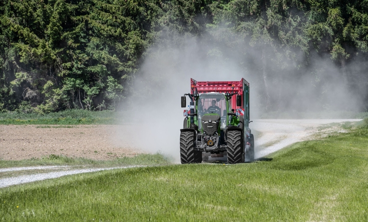 Ein Fendt 724 Vario Gen7.1 transportiert einen Strautmann Ladewagen auf dem Feldweg. Ein grüner Fendt 724 Vario Gen7.1 fährt im Gespann mit einem Strautmann Ladewagen über einen Feldweg direkt auf die Kamera zu. Im Hintergrund ist ein Wald zu sehen.