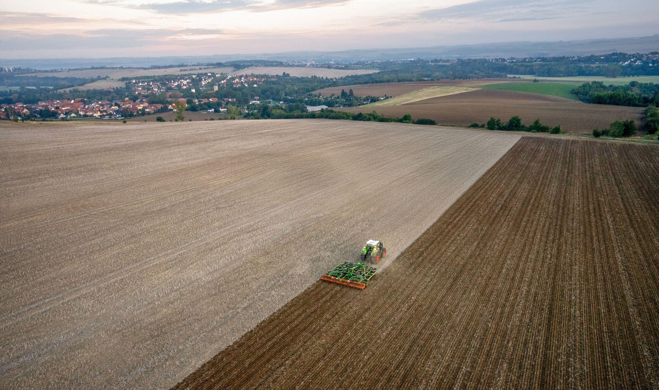 Ein grüner Fendt 1000 Vario bearbeitet mit einem Grubber ein großes Stoppelfeld. Die Vogelperspektive zeigt weitere Flächen und ein Dorf im Hintergrund bei Morgenstimmung.